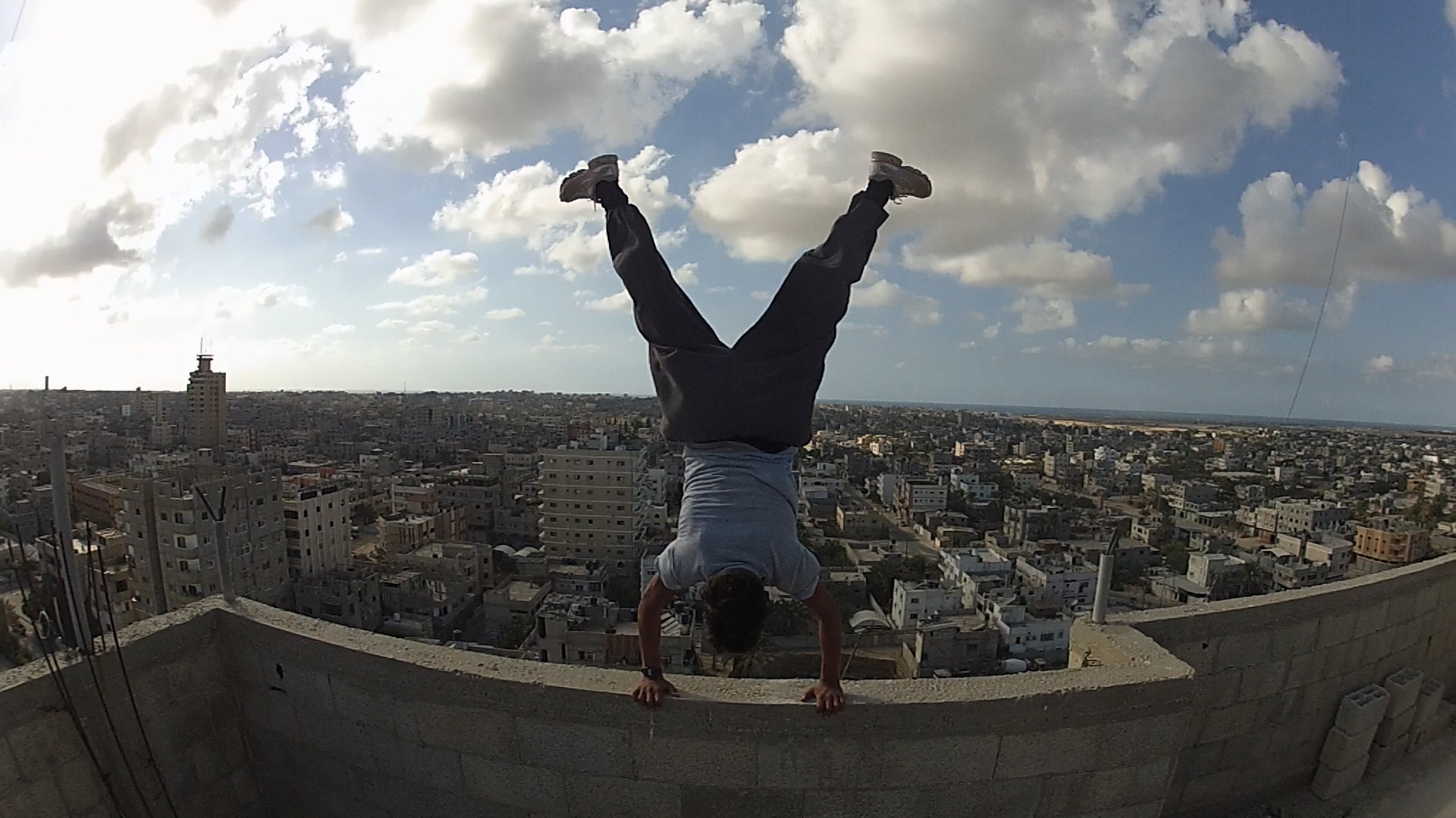 A person performs a handstand on a rooftop overlooking a sprawling cityscape under a partly cloudy sky.