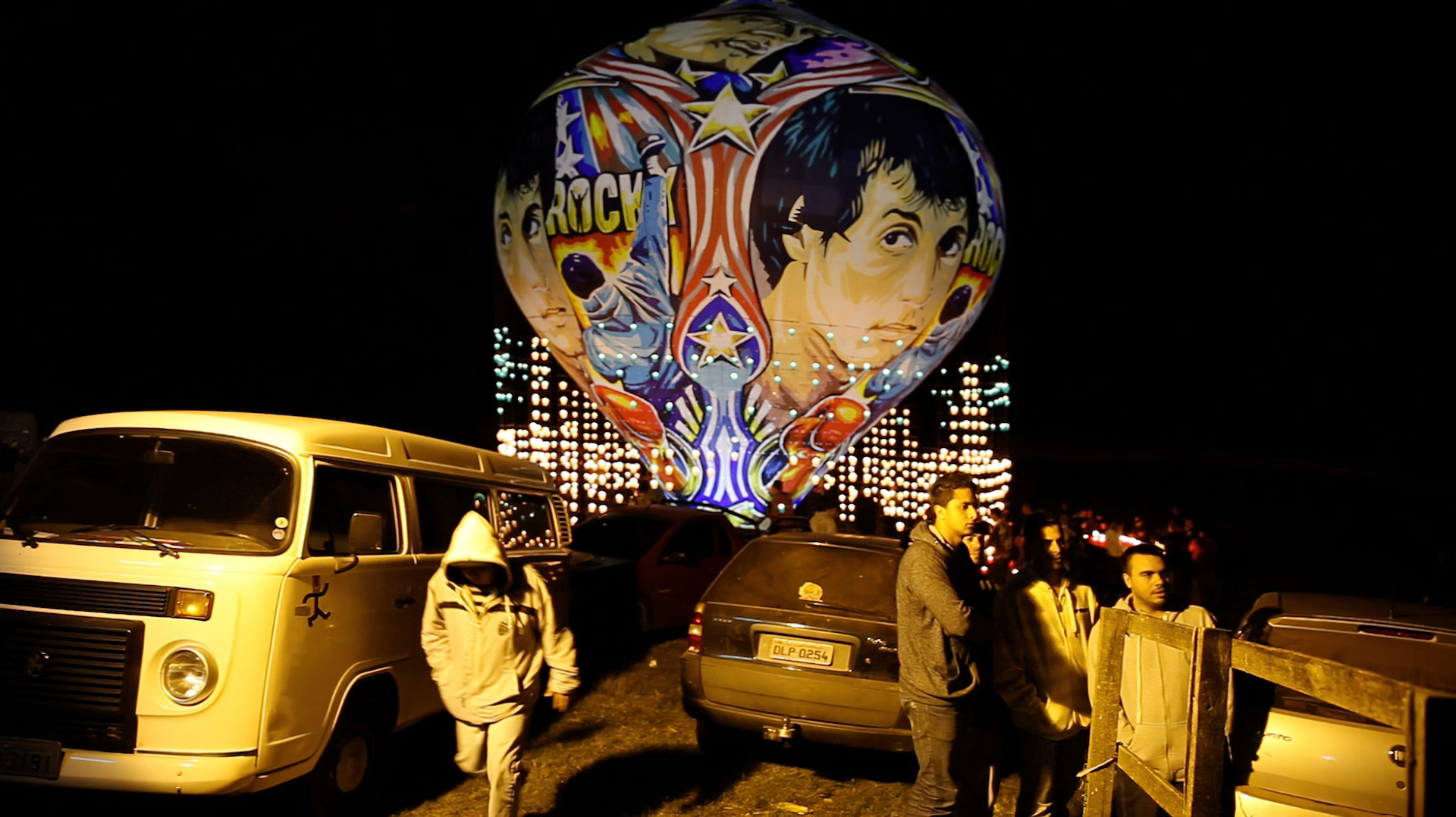 A vibrant hot air balloon glows against the night sky, surrounded by parked cars and people enjoying the festive atmosphere.
