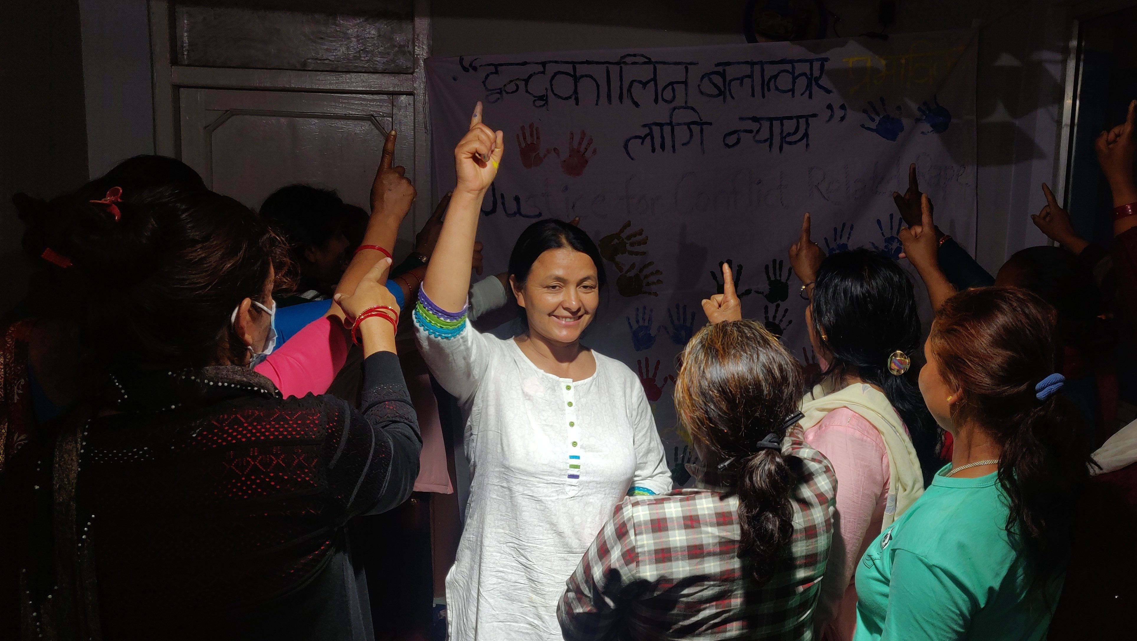 Een groep vrouwen verzamelt zich en steekt eenstemmig hun wijsvingers op. In het midden leidt een glimlachende Devi Khadka het gebaar. Achter hen is een spandoek met Nepalese tekst en kleurrijke handafdrukken te zien.