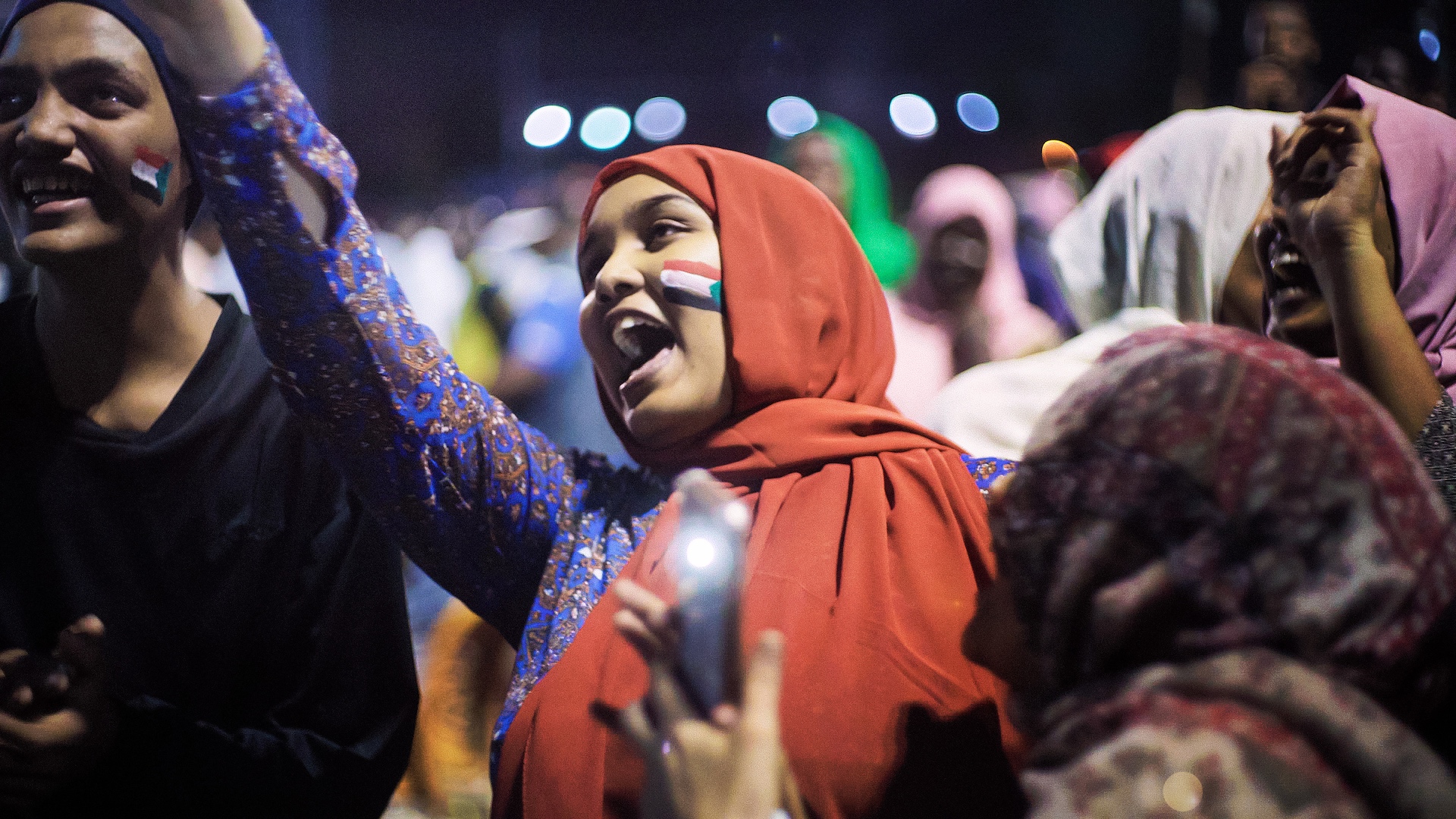 A group of people with the Sudanese flag on their cheeks stand together, some raising their hands and others holding mobile phones in a lively night atmosphere.