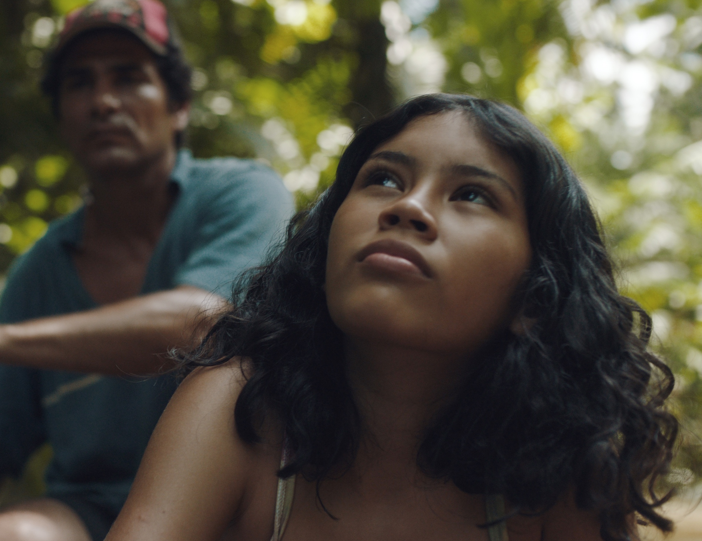 A young girl with curly hair sits in a lush green jungle, while a man in the background appears to be guiding her on a journey.