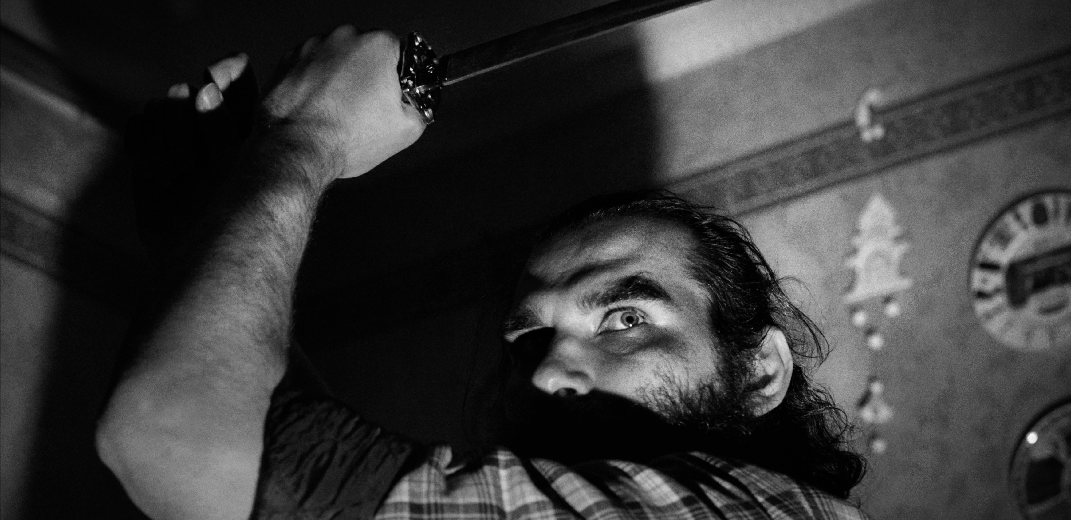 A man raises a katana sword overhead, captured in dramatic black and white, with a shadowy background suggesting tension.