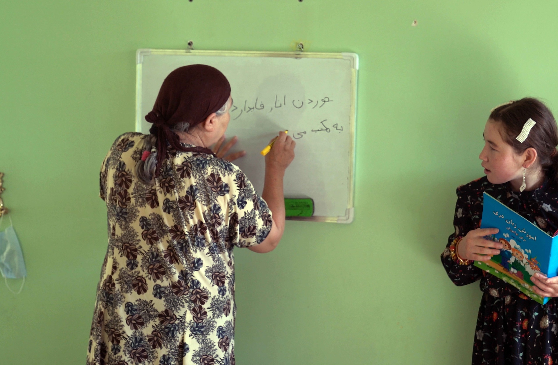 An elderly woman writes on a whiteboard in a classroom while a young girl holds a colorful book. The wall is painted green.