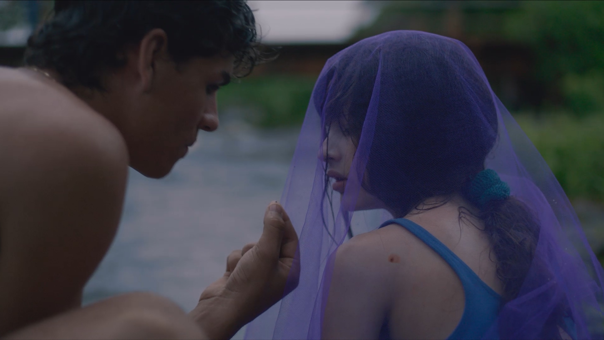 A person gestures towards a young girl, whose head is covered with a purple veil, as they sit by a waterbody in a lush, green setting.