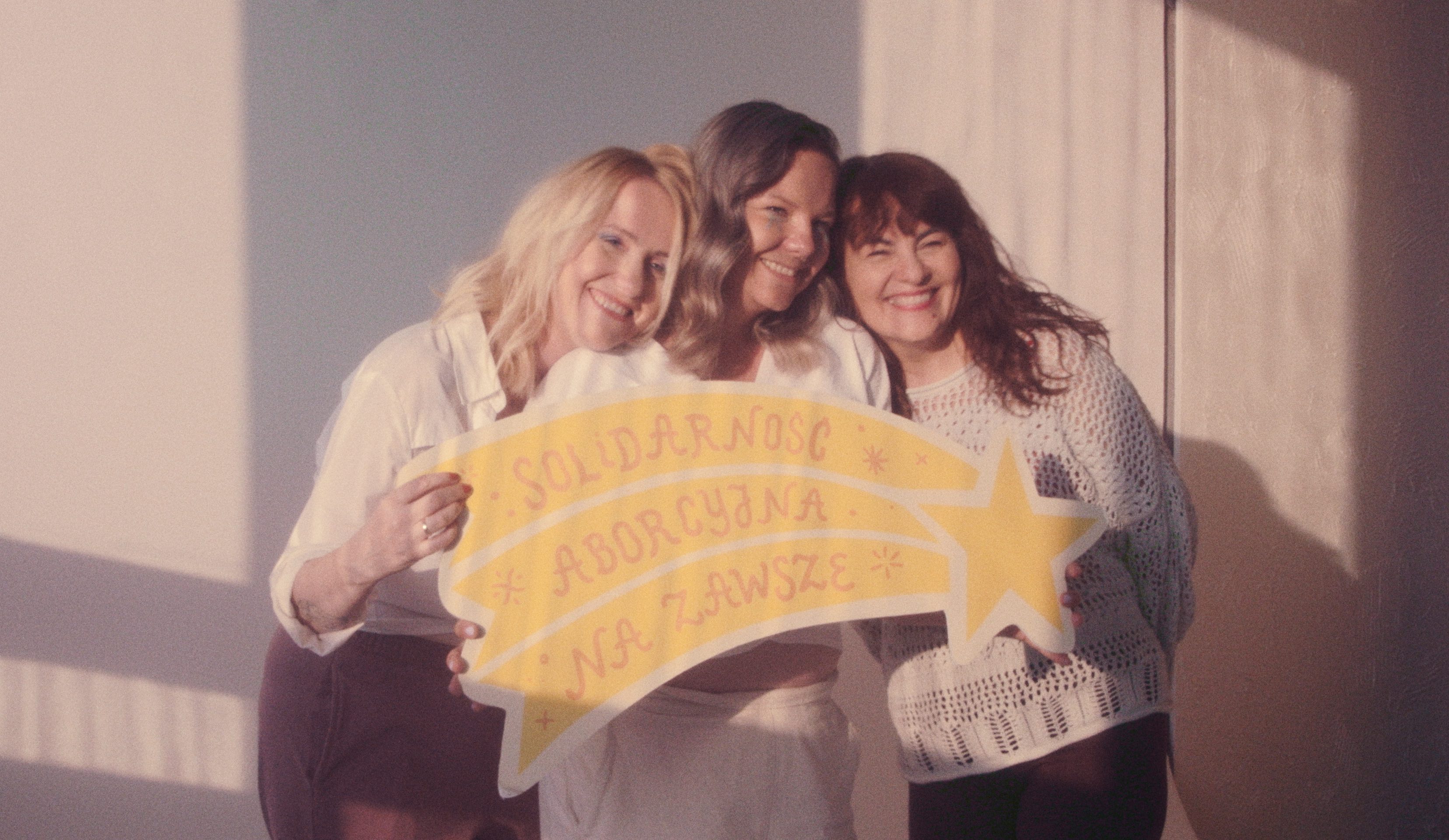 Three smiling women stand close together, holding a yellow sign shaped like a shooting star. The sign displays the Polish phrase "Solidarność Aborcyjna Na Zawsze," which translates to "Abortion Solidarity Forever."