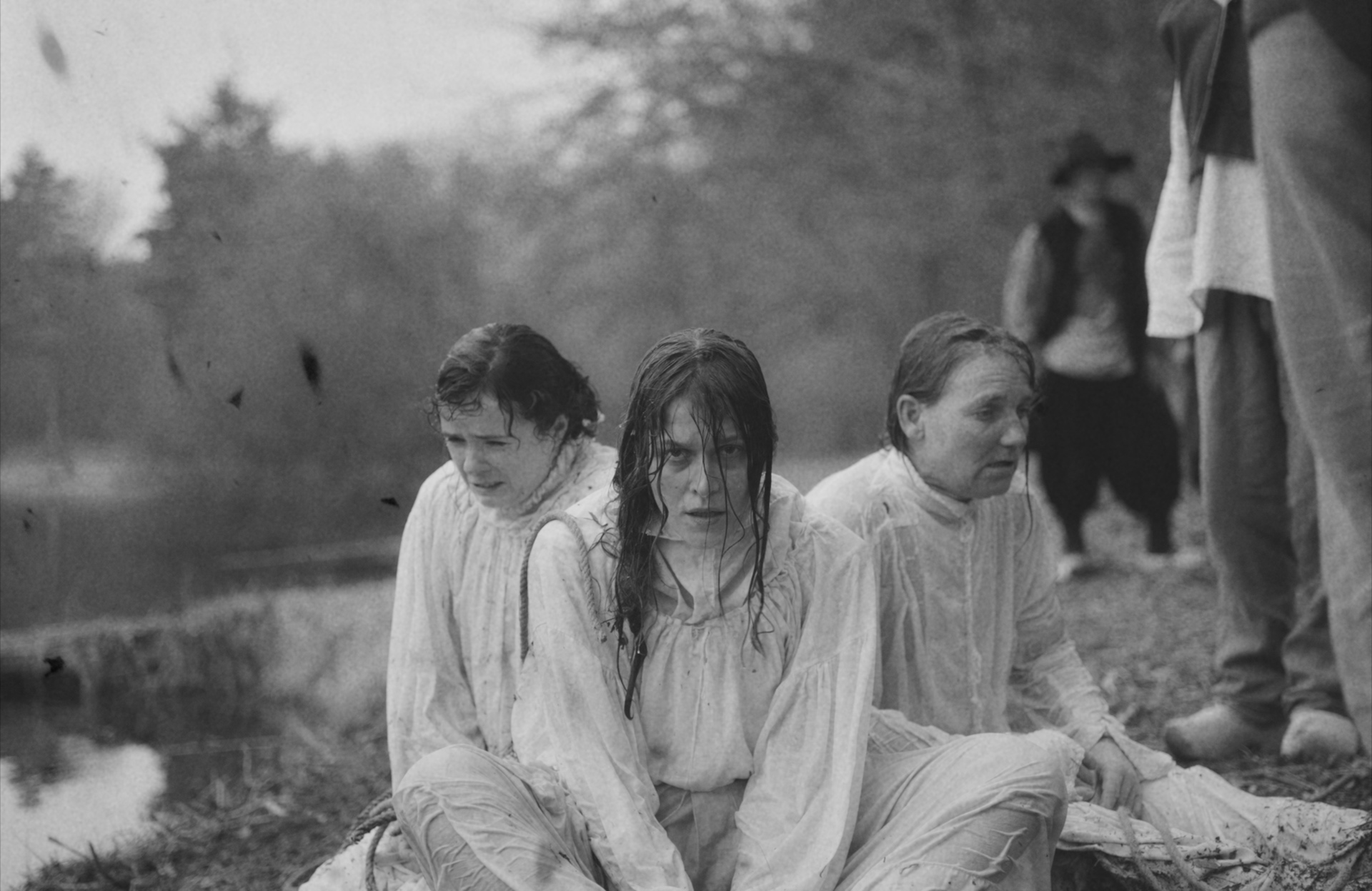 Drie vrouwen in natte, witte kleding zitten bij een waterpartij, omringd door bomen en toeschouwers in historische kledij.