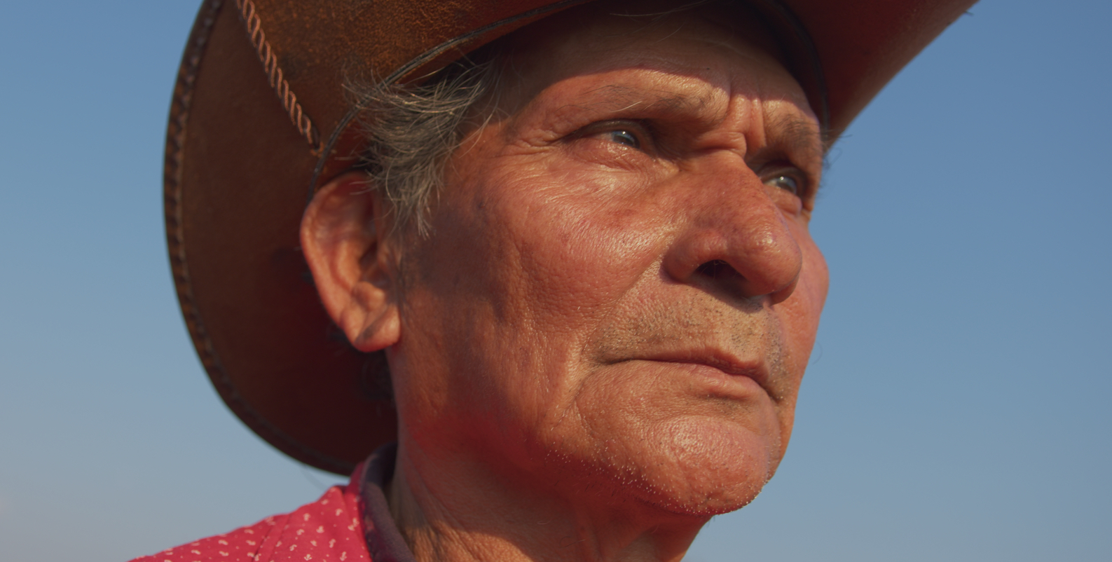 A man wearing a wide-brimmed hat and a red polka-dot shirt stands against a clear blue sky.