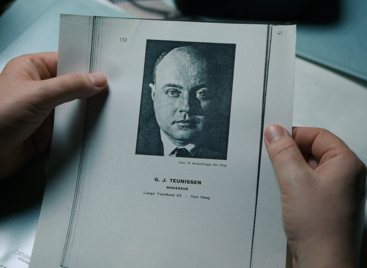 A person holds a document featuring a black and white portrait image along with the name G. J. Teunissen and an address in Den Haag.