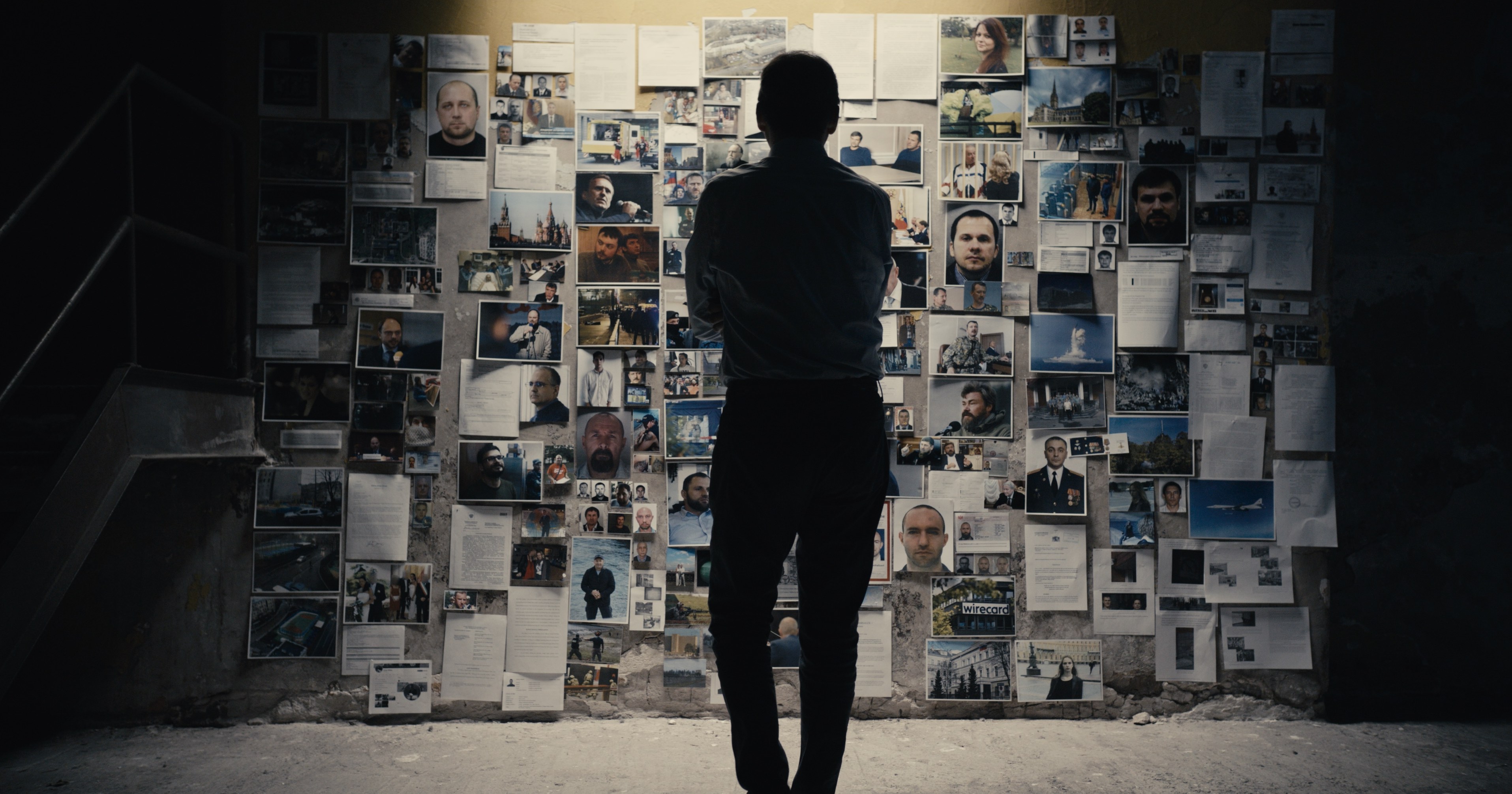 A silhouette of a man stands facing a wall covered with various photos and documents in a dimly lit room.