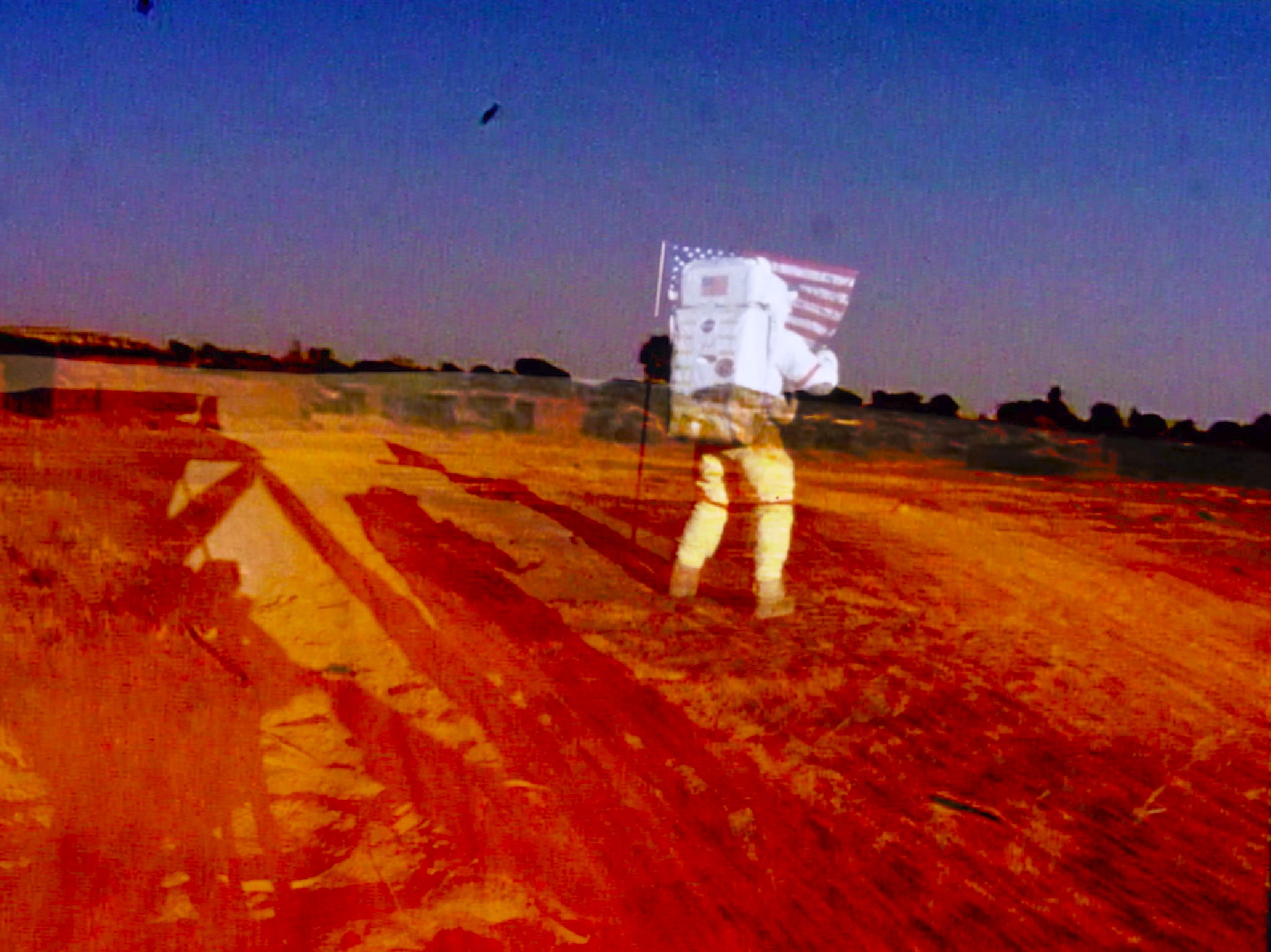 Een astronaut in een ruimtepak loopt over een stoffig oppervlak en houdt een Amerikaanse vlag vast tegen een strakblauwe lucht