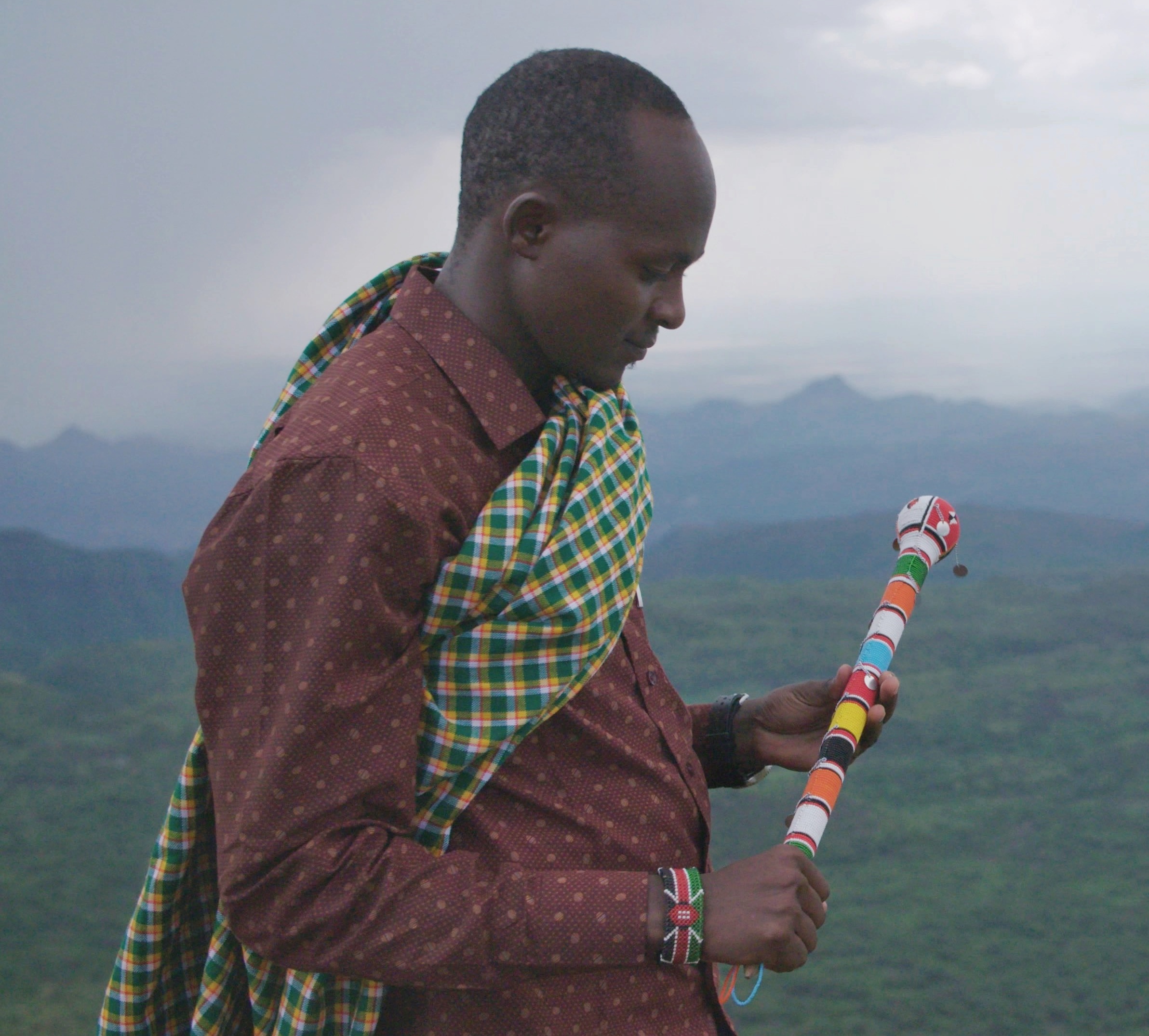 Man holding a colorful staff, wearing a patterned shirt and a checkered cloth over his shoulder, stands outdoors with a hilly landscape and cloudy sky in the background.