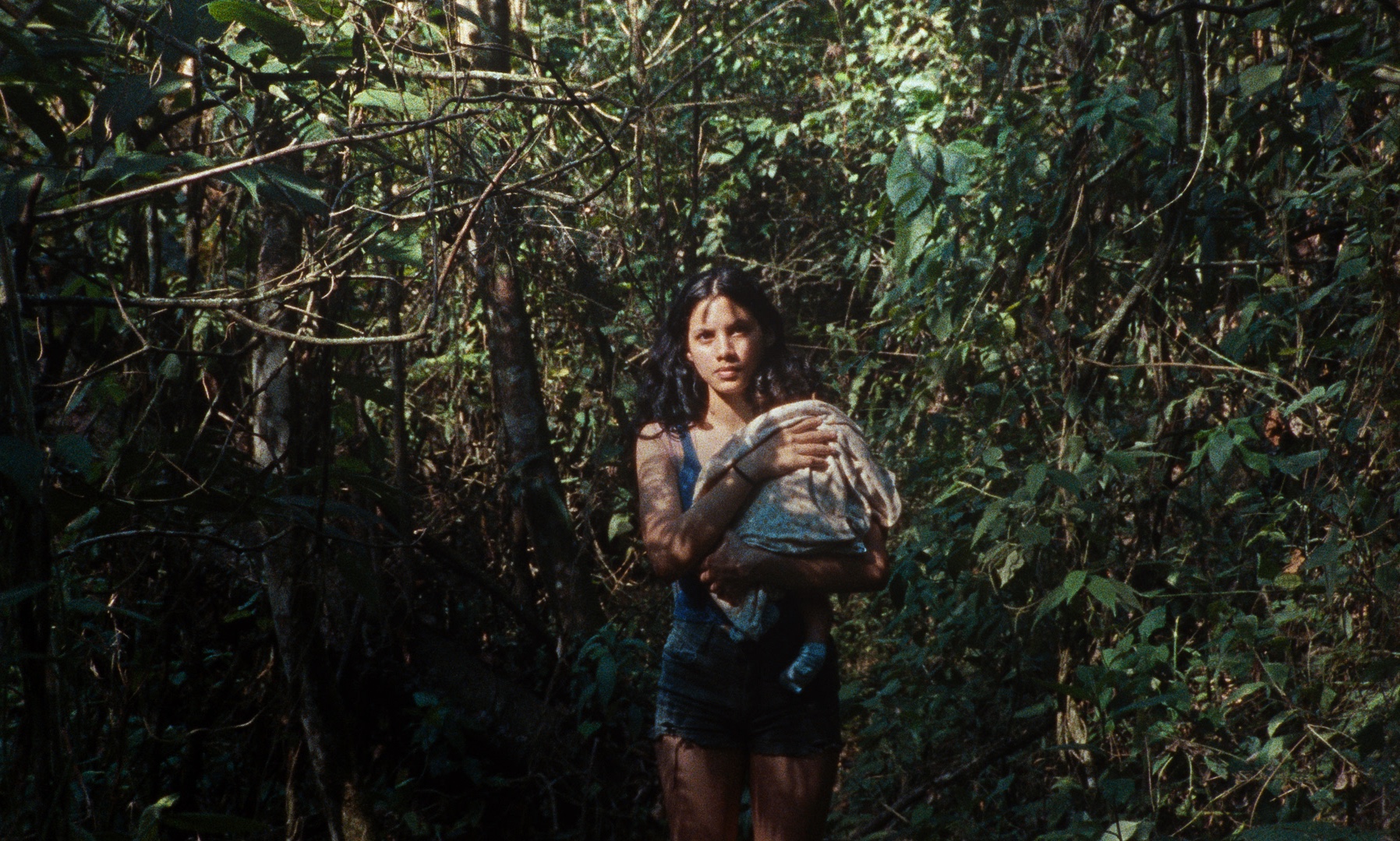 A woman holding a baby stands amid dense greenery in a forest