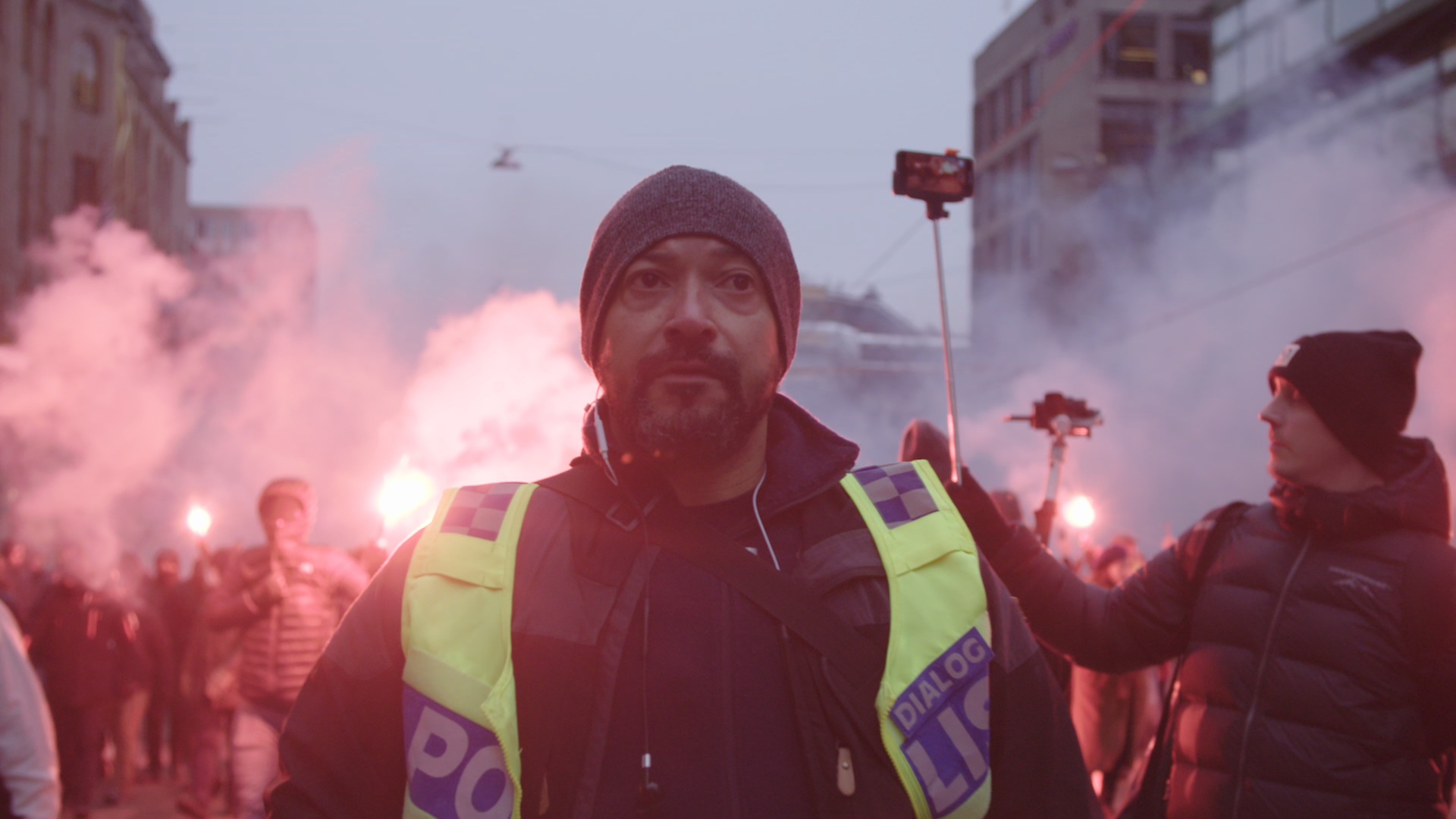 A policeman in a yellow vest stands among protesters carrying red smoke torches.