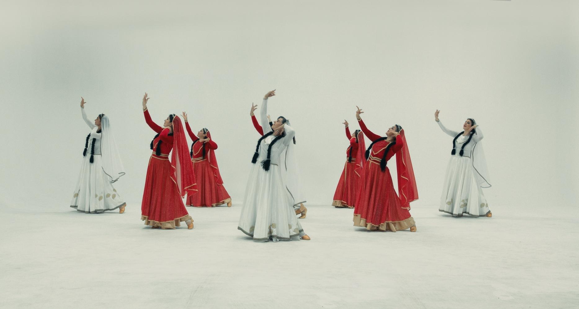 A group of female dancers in vibrant red and white traditional costumes perform gracefully against a bright, minimalist backdrop.